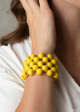 Load image into Gallery viewer, An oversized collection of sunny yellow wooden beads are threaded along stretchy bands that ornately weave across the wrist, creating a decorative summery pattern.
