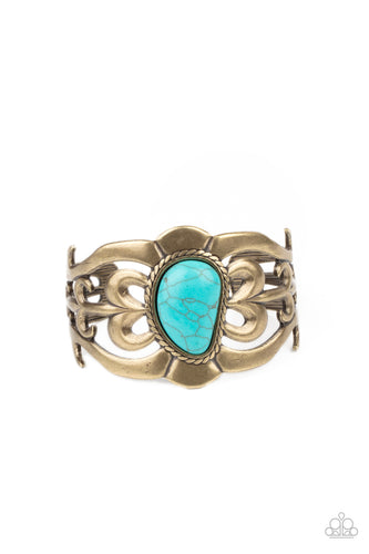 An asymmetrical turquoise stone is pressed into the center of a brass cuff layered with filigree patterns around the wrist for a rustic flair.