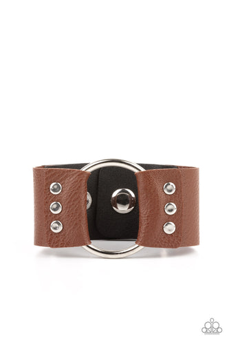 An oversized silver hoop is studded in place between the center of studded pieces of black and brown leather, creating a rustic centerpiece around the wrist. Features an adjustable snap closure