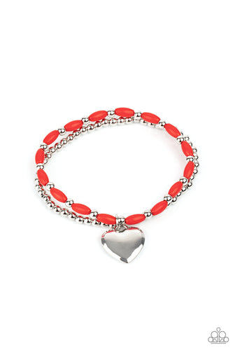 A shiny silver heart dangles from a strand of fiery red beads. It is paired with a strand of round silver beads threaded along a stretchy band for a whimsical display around the wrist.