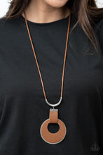 Load image into Gallery viewer, A large cutout silver disc, overlaid with brown leather, is cradled in a wide brown leather strap. The luxe pendant sways from a U-shaped silver cylinder threaded along the end of a lengthened polished brown cord for an indulgently lavish finish. Features a sliding bead closure. 
