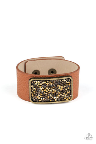 A collision of topaz and aurum rhinestones collect inside a brass rectangular frame that is studded in place in the center of a thick brown leather band, creating a sparkling statement piece around the wrist. Features an adjustable snap closure.