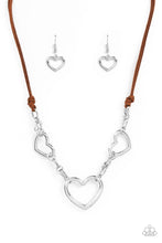 Load image into Gallery viewer, Strands of brown suede knot around sections of chunky silver chains that have been adorned in oversized silver heart frames, resulting in a flirtatious display below the collar. Features an adjustable clasp closure.
