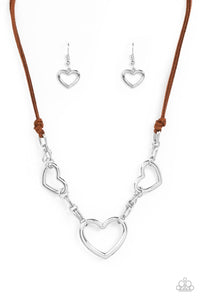 Strands of brown suede knot around sections of chunky silver chains that have been adorned in oversized silver heart frames, resulting in a flirtatious display below the collar. Features an adjustable clasp closure.