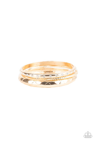 A thick gold bangle that is embossed and faceted in an edgy geometric pattern joins a mismatched pair of white rhinestone encrusted and diamond cut gold bangles that glide along the wrist, displaying a gritty pop of shimmer. 