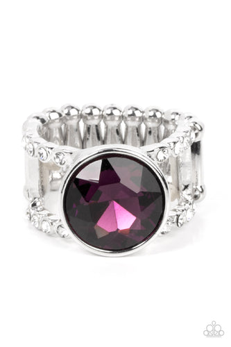 A dramatically oversized brilliant purple solitaire gem, encased in a simple silver frame, sits atop rhinestone encrusted layered bands creating an unapologetically head-turning statement. Features a stretchy band for a flexible fit.