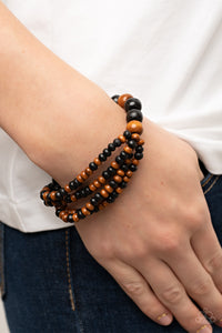 Stretchy strands of dainty brown and black wooden beads attach to a single strand of oversized brown and black wooden beads, resulting in colorful layers around the wrist.