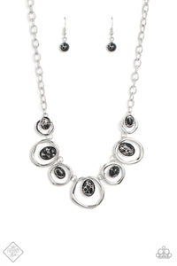 Marbled black stones are encased in classic silver frames and attached to the inside edges of warped silver hoops. The haphazard placement of the stones and the irregular shape of the silver rings add a handcrafted touch to the design, as the asymmetrical frames fall along the collar at the end of a thick silver chain. Features an adjustable clasp closure. As the stone elements in this piece are natural, some color variation is normal.
