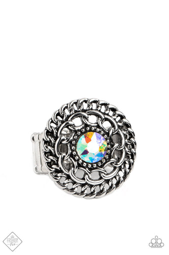 An iridescent rhinestone with orange undertones glimmers at the center of a frame of radiating chain links. Tiny silver studs wrap closely around the sparkly centerpiece, adding extra texture to the design. Features a stretchy band for a flexible fit. Due to its prismatic palette, color may vary.  Sold as one individual ring.