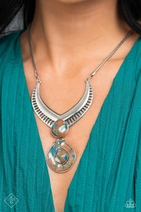 A thick, silver, v-shaped frame, with airy, vented cutouts, sits boldly at the end of a silver snake chain. A widened teardrop-shaped stone, marbled with swirls of blue, white, and brown, sits at the center of the dramatic frame, adding a pop of color to the regal design. A second teardrop-shaped stone — with a third, smaller stone nestled within a silver border at its center — swings from the polished stone above, adding eye-catching movement and intriguing detail. 