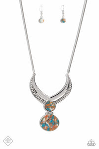 A thick, silver, v-shaped frame, with airy, vented cutouts, sits boldly at the end of a silver snake chain. A widened teardrop-shaped stone, marbled with swirls of blue, white, and brown, sits at the center of the dramatic frame, adding a pop of color to the regal design. A second teardrop-shaped stone — with a third, smaller stone nestled within a silver border at its center — swings from the polished stone above, adding eye-catching movement and intriguing detail. 