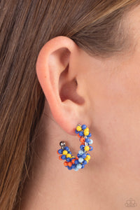 A colorful collection of royal blue, yellow, light blue, and orange seed beads are wrapped along the outside curve of a shiny, silver hoop by shimmery silver wire for a handcrafted, carefree finish. A shiny silver ball is affixed to the end of the hoop, reminiscent of a barbell fitting. Earring attaches to a standard post fitting. Hoop measures approximately 1" in diameter. 